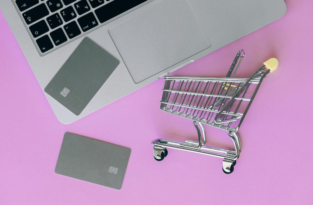 A miniature shopping cart, credit cards, and a laptop against a pink bankground