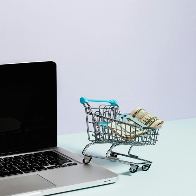 A mini shopping cart filled with cash, next to a laptop