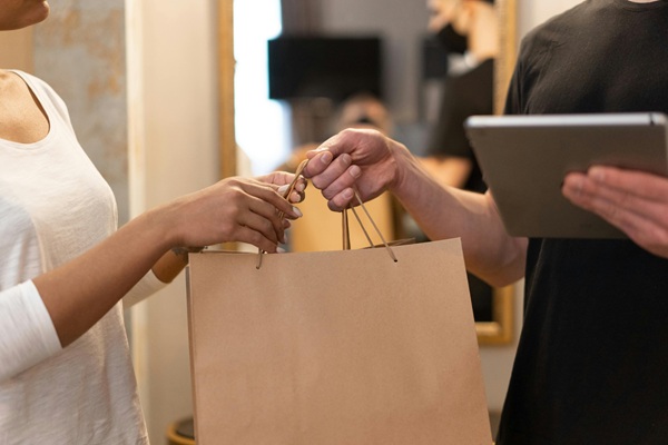 A person handing over a shopping bag to another person holding a tablet